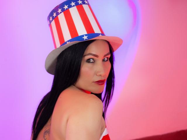 VictoriaEloit with long black hair, wearing a red and white striped top hat, red choker, and looking back over her shoulder, showing her tattoo and red lipstick.