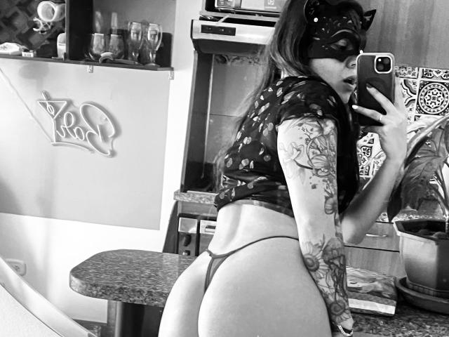 Black-and-white selfie of SujannaVarma with a cat mask, tattooed arm, and thong, showing her round ass, wearing a dark floral top, holding a phone, in a kitchen.