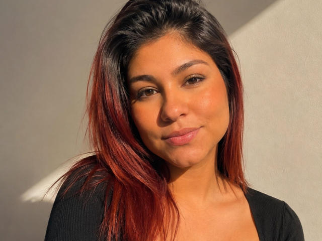 SamanthaBlossom a les cheveux noirs avec des mèches rouges, des yeux marrons et un léger sourire. Elle porte un haut noir, et la lumière du soleil met en valeur sa peau bronzée et son maquillage discret..