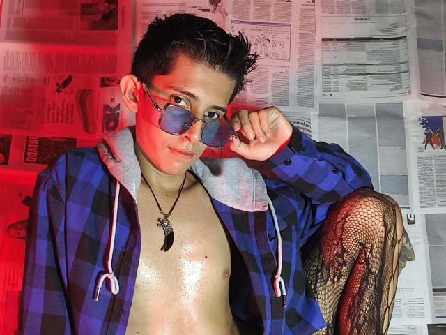 GomitaBay mit kurzen, dunklen Haaren, trägt eine blaue, karierte Kapuzenjacke, eine Brille und Fischernetzstrümpfe, ist obenhin bekleidet, zeigt eine glänzende Brust, hält eine Brille und eine Kreuzkette, rotes Licht im Gesicht..