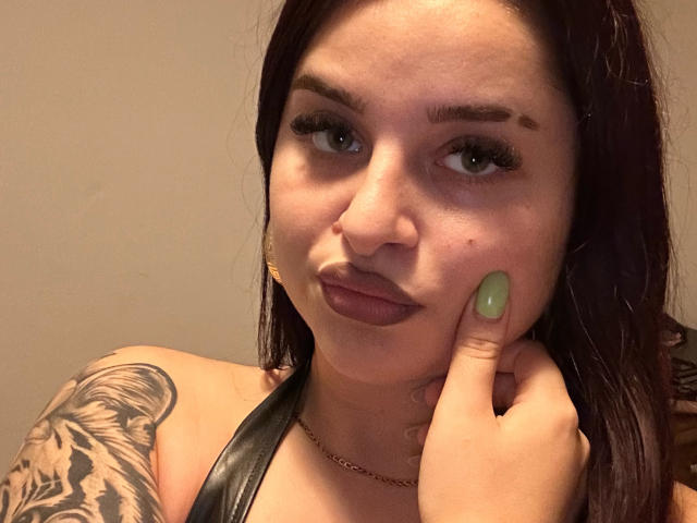 Luucie, avec des ongles verts, des cheveux brun foncé et un tatouage sur l'épaule gauche, se tient la joue, porte un haut noir à encolure dégagée et regarde l'appareil photo avec un léger froncement de sourcils..