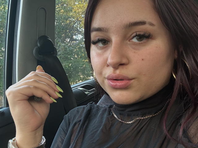 Luucie with green nails, puffy pink lips, and a black sheer top, wearing a chain, in a car, looking at the camera with a slight pout.