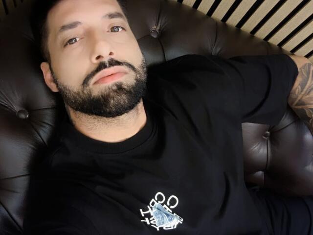 TommyDum with a beard, black shirt, and tattooed arm, lying on a brown leather couch, looking at the camera with blue eyes and a slightly serious expression.