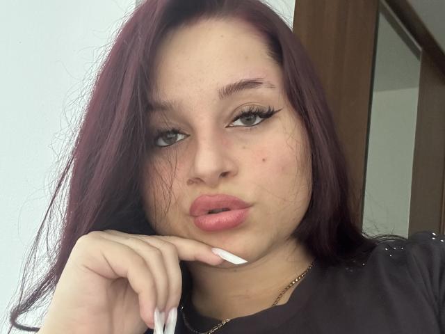 Luucie with dark purple hair, pouty pink lips, and thick black eyeliner, touching her chin with white-manicured nails, looking seductively at the camera.