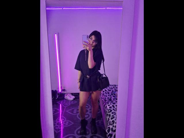 PaulethJoens in a black shirt dress, holding phone, purple neon, standing, black boots, leopard bed, black bag, mirror selfie, long dark hair, purple light, patterned floor.