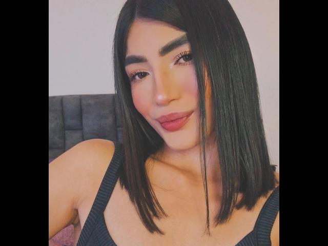 PaulethJoens with straight black bob, thick eyebrows, pink blush, red lipstick, and black tank top, showing her chest. Her tits are small and her skin is tan.