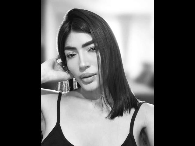 Black and white photo of PaulethJoens with straight dark hair, looking at camera, hand on head, wearing a spaghetti strap top, showing small tits, full lips, and dark eyebrows.
