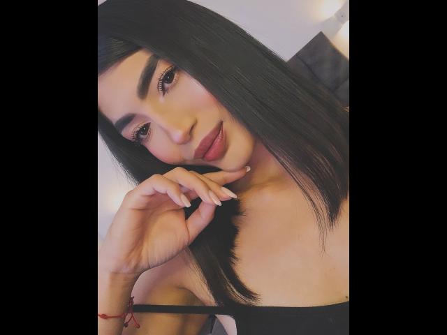 PaulethJoens with black hair, full eyebrows, and pink lipstick, touching her chin with a finger, wearing a black strap, smiling subtly, in a soft-lit photo.