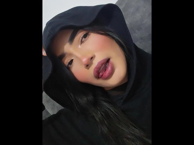 PaulethJoens with dark lipstick, black hood, and long black hair, eyes half-open, slightly parted red lips, looking seductive, wearing a black hoodie, soft light on face.