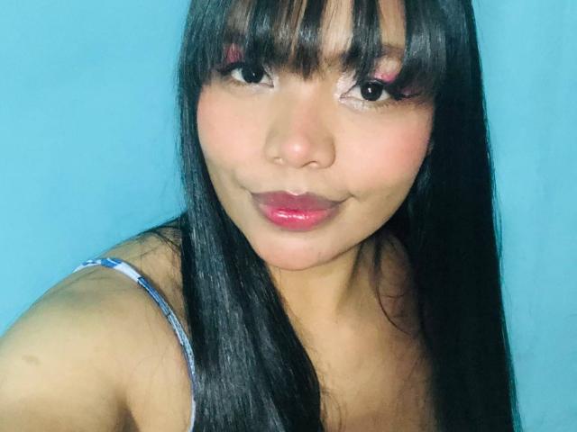 Kelly Jackson mit glänzendem, schwarzem Haar, rosafarbenen Augenlidern und glänzenden, rosafarbenen Lippen, trägt ein blaues Top mit Spaghetti-Trägern und lächelt leicht. Ihr süßes Gesicht und ihre glatte Haut sind sichtbar..