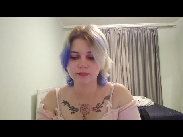PaolaStohler with blue and blonde hair, pink off-shoulder top, chest tattoos, looking down, slight pout, white walls, grey curtains, bed with dark blanket in background.