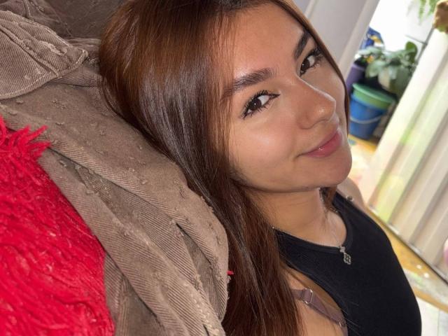 AlessiaFerry with long brown hair, brown eyes, and a small smile, wearing a black top, leaning on a brown textured jacket with a red fabric.