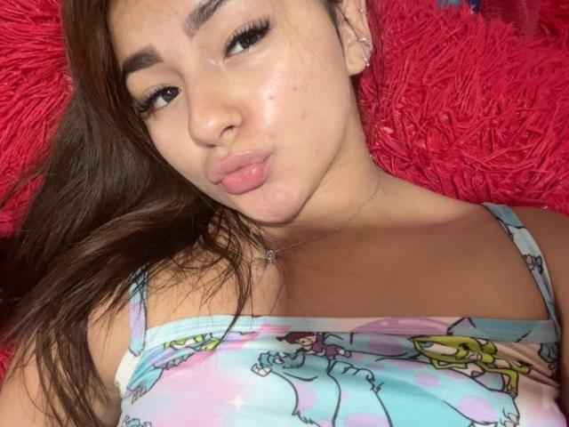 AlessiaFerry with pouty lips, brown hair, wearing a colorful, cartoon-print dress, lying on a red, fluffy pillow, showing her shiny, slightly freckled face and small, pointed nose.