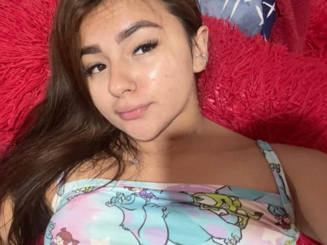 AlessiaFerry with long brown hair, subtle makeup, and a slight smile, wearing a colorful floral top, lying on a red, fluffy surface. Boobs visible, small earrings in.