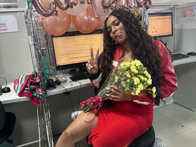 KathePalmer, with long black curly hair, red dress, and red jacket, sits in office, flashing peace sign, holding yellow flower bouquet, red ribbon on thigh. Balloons overhead.