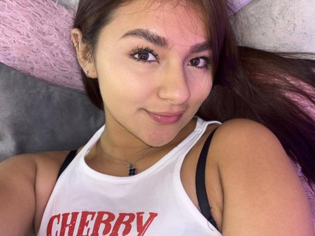 AlessiaFerry with brown hair, light skin, and a small nose, wearing a white tank top with red "CHERRY" text, black straps, and a necklace, smiling.