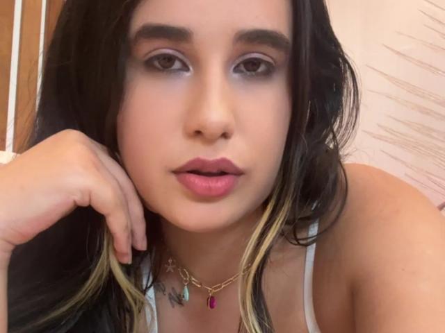 KatieDesirX, avec des cheveux foncés, des yeux marron clair, du rouge à lèvres rose, portant un haut blanc, un collier en or avec un bijou rose, la main reposant sur la joue, un tatouage visible sur la poitrine..
