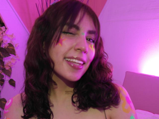 AshlyValdes, avec ses cheveux bruns ondulés, clignant de l'œil, souriant, avec du maquillage coloré sur le visage, un éclairage rose et violet, des tatouages sur l'épaule, un lit blanc en arrière-plan, un mur violet. Seins partiellement visibles, nue..