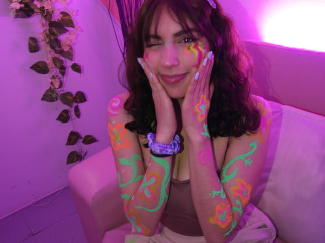 AshlyValdes a du body painting fluorescent sur ses seins, ses fesses et ses bras, elle fait un clin d'œil en posant ses mains sur ses joues, dans une pièce éclairée en violet, et elle porte un élastique pour les cheveux violet et un haut marron..