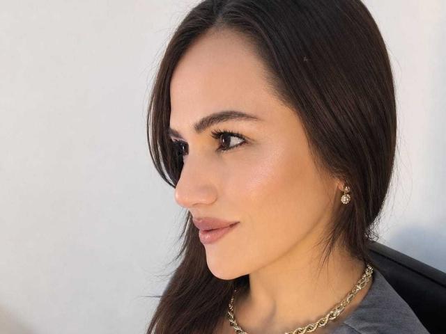 CameronViviane tiene el pelo largo y castaño, ojos marrones, y lleva un collar de cadena de oro con pequeños pendientes. Está mirando hacia un lado con una sonrisa sutil..