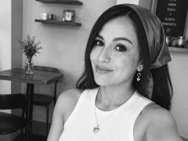 Selfie en blanco y negro de CameronViviane con un pañuelo en la cabeza, aretes con forma de corazón y una camiseta blanca, mostrando una sonrisa encantadora, un pequeño aro en la nariz y un collar con forma de corazón. Los senos no son visibles..