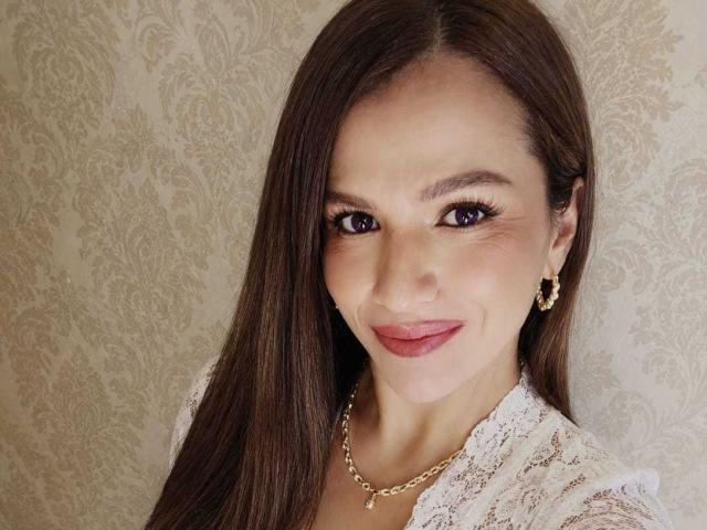 CameronViviane tiene el pelo largo y castaño, ojos morados, y está sonriendo con lápiz labial rosa. Lleva una blusa de encaje, un collar dorado y aretes de perlas. No se ven los senos..
