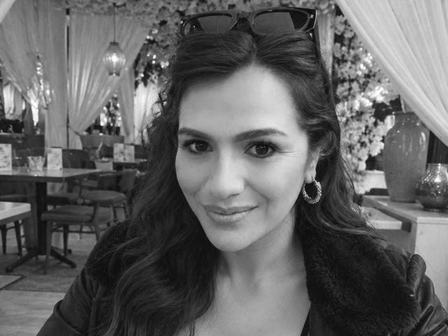 CameronViviane tiene el cabello largo y ondulado, lleva gafas de sol oscuras en la cabeza, aros grandes y una chaqueta de cuero, con una sonrisa sutil, en una fotografía en blanco y negro..