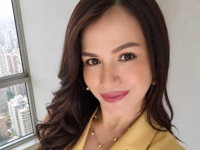 CameronViviane tiene el cabello castaño ondulado, ojos marrones, lápiz labial rosa, una camisa amarilla, aretes dorados y un collar. Su trasero no es visible. Está sonriendo a la cámara..