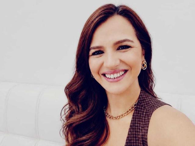 CameronViviane tiene el pelo largo y castaño, una sonrisa encantadora, y lleva una blusa marrón a cuadros con aretes dorados y un collar de cadena, luciendo atractiva sobre un fondo blanco..