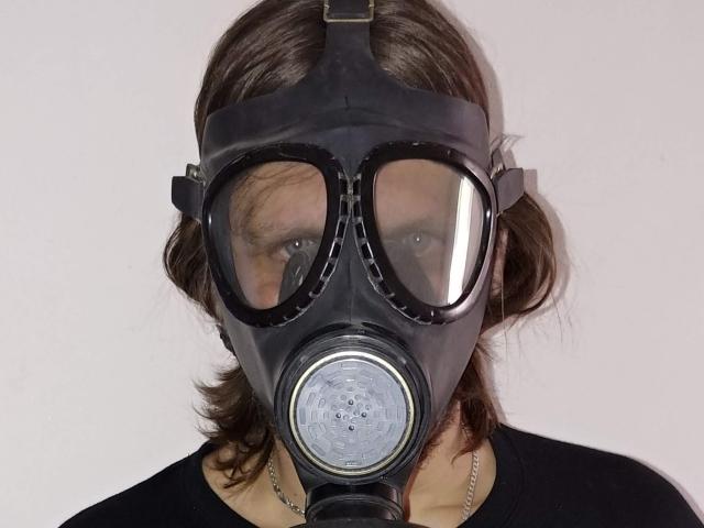 FrancimooR usando uma máscara de gás preta com olhos grandes e transparentes e um filtro de metal, tem cabelo castanho ondulado e está vestindo uma camisa preta..