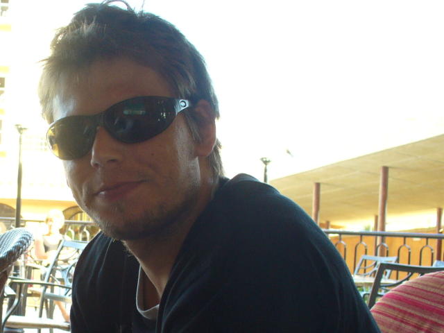 FrancimooR, com óculos de sol pretos, cabelo castanho curto e uma camisa preta, exibe um leve sorriso, tem uma barba discreta e uma camisa branca por baixo..
