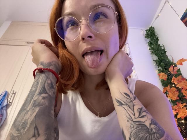 Sarah Rousen, com cabelo ruivo, óculos e língua de fora, vestindo uma camiseta branca, mostrando os braços tatuados e um bracelete vermelho, em um selfie..