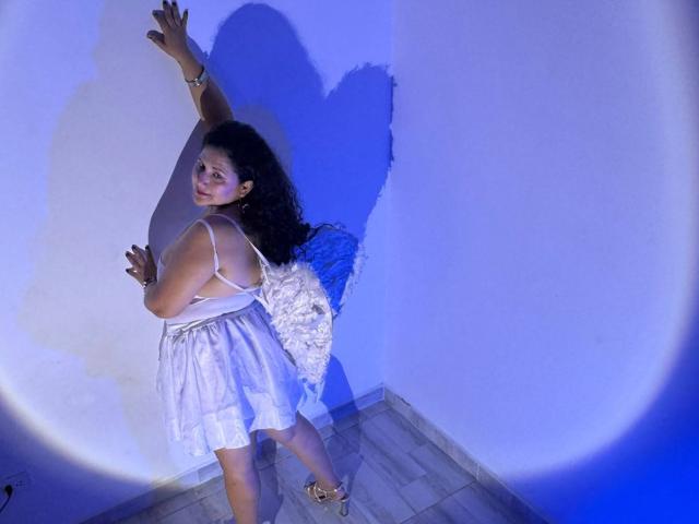 GenesisMontenegro mit lockigem, dunklem Haar, in einem weißen Kleid, steht an einer Wand, die Arme erhoben, ein Schatten an der Wand, blaues Licht, Holzboden, blickt über die Schulter, lächelnd..