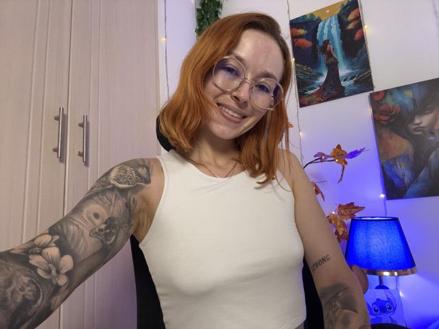 SarahRousen with red hair, glasses, white sleeveless top, showing tattooed arms, smiling, in a room with a blue lamp, colorful wall art, and a white wardrobe.