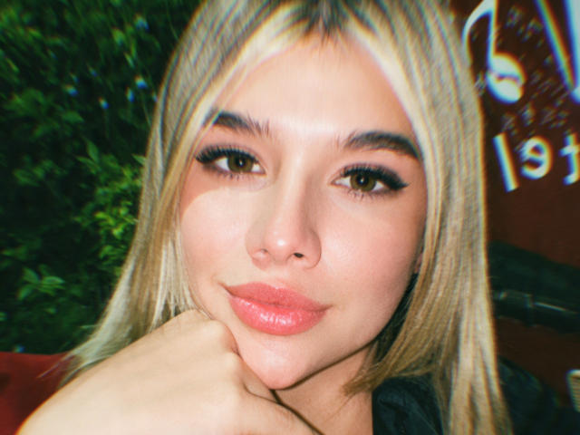 SashaPolet heeft blond haar, dikke wenkbrauwen en ze frumelt, waarbij haar kin op haar hand rust, en ze laat haar glanzende, roze lippen zien. Ze kijkt recht in de camera..