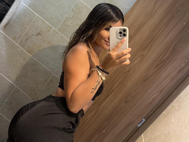 SashaPolet with wet black hair, wearing a black sports bra and high-waisted black pants, taking a mirror selfie with an iPhone, showing her curvy ass and small tits.