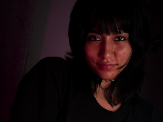 JenFoxy, con cabello negro oscuro, vestida con una camisa negra y un collar, tiene una sonrisa sutil y un rostro ligeramente sonrojado, en una iluminación púrpura tenue, mostrando su pequeño y lindo trasero..