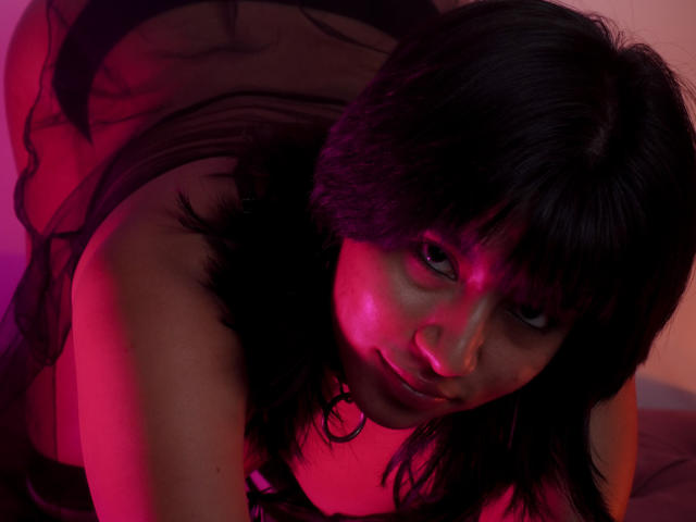 JenyFoxy steht auf allen Vieren, trägt ein durchsichtiges schwarzes Oberteil, und ihr Gesicht ist von rotem und violettem Licht beleuchtet. Sie hat dunkles, welliges Haar. Ihre Brüste sind leicht sichtbar..