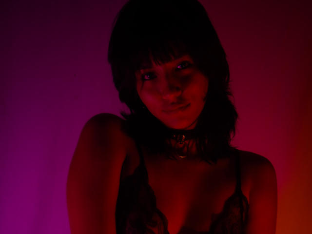 JenyFoxy, con cabello oscuro, vistiendo un sujetador de encaje negro y un collar, iluminada con luces rojas y moradas, mostrando sus senos y una sonrisa sutil..