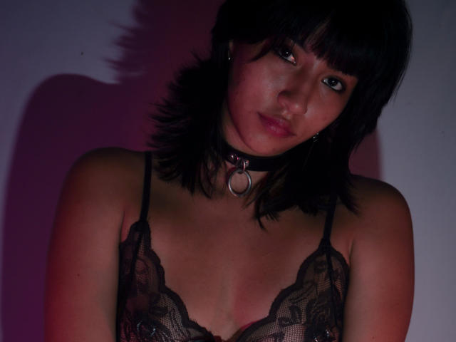 JenyFoxy con lingerie in pizzo nero, collana con anello, e capelli scuri corti, che mostra piccoli seni, capezzoli rosa e un'espressione seria in una luce fioca..