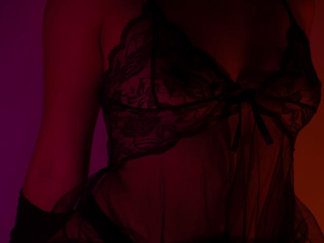 La lencería rojo oscuro de JenyFoxy, con encaje, muestra sus pechos, bajo una iluminación suave en tonos púrpura y naranja, creando un efecto sensual y sombrío. Su brazo es parcialmente visible..