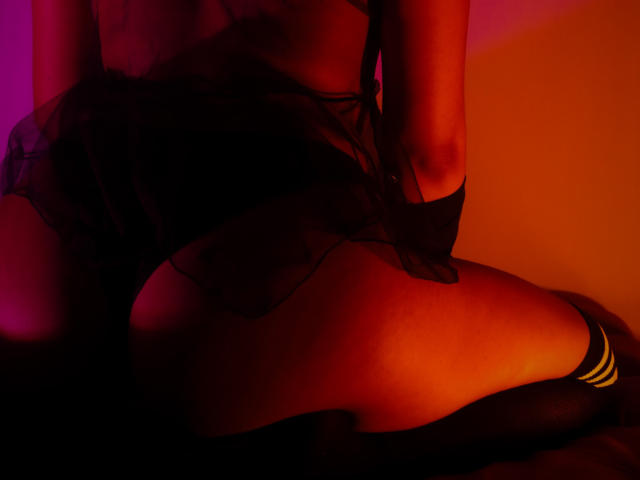JenyFoxys nackter Hintern, bedeckt mit einem transparenten schwarzen Stoff, wird durch rotes und oranges Licht hervorgehoben. Sie trägt schwarze, hohe Socken mit gelben Streifen. Ihre Hände liegen auf ihren Oberschenkeln..