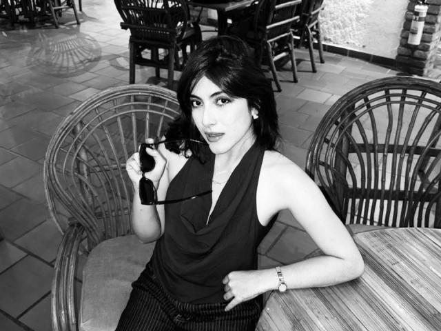 SpencerLe, avec des cheveux foncés, tenant des lunettes, portant un débardeur noir, assise dans une chaise en osier, affichant un léger sourire, portant un bracelet, sur une photo en noir et blanc..