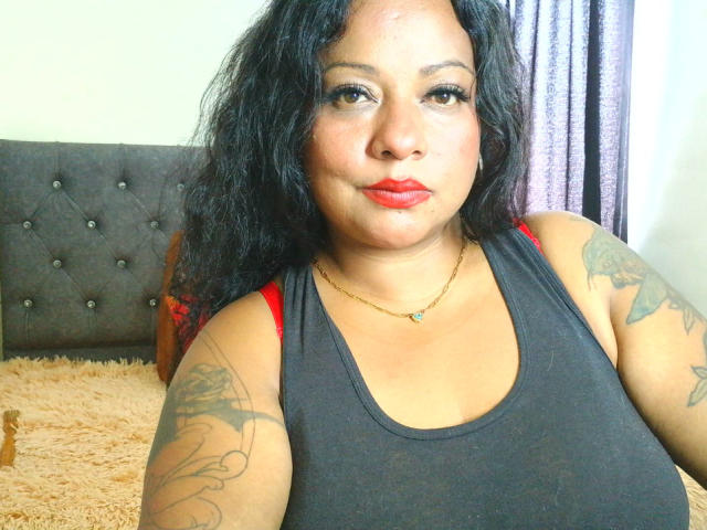 CurvySensation mit lockigem, schwarzem Haar, rotem Lippenstift, schwarzem Tanktop, Tattoos an den Armen, einer goldenen Halskette und roten BH-Trägern, sitzt auf einem Bett mit einem grauen, gepolsterten Kopfteil..