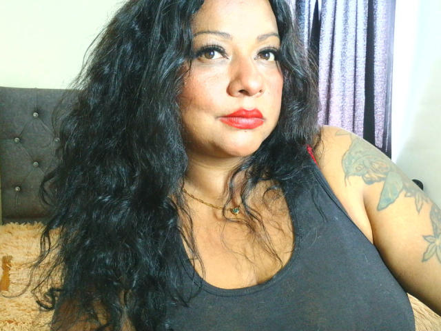 CurvySensation met krullend zwart haar, rode lippenstift, tatoeages op haar rechterarm, gekleed in een zwart tanktopje, zittend op een bed, kijkend naar de camera..