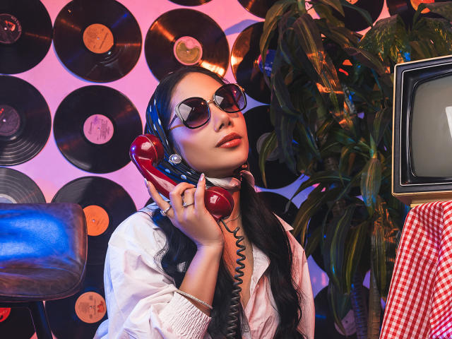 ArianaRogers with red corded phone, black hair, sunglasses, white shirt, holding phone to ear, looking up, plant on right, vintage TV on left, red checkered cloth on bottom.