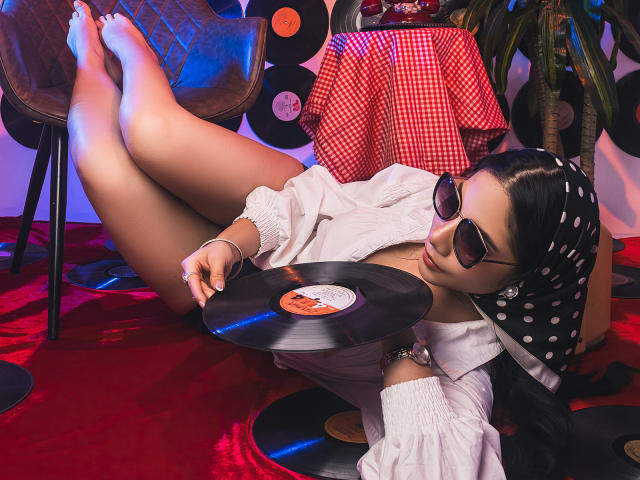 ArianaRogers with dark hair, polka dot headscarf, sunglasses, white shirt, lying on red floor, legs up, holding vinyl record, red checkered cloth, vinyl records in background.