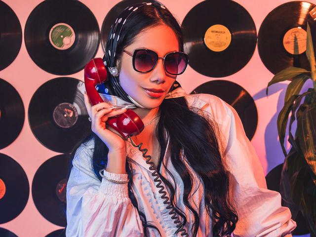 ArianaRogers with long black hair, wearing sunglasses, a white jacket, and a red phone, holding it to her ear, looking sexy. Vinyl records in the background.