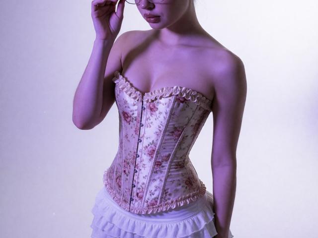Destinis with a floral, pink-ribboned corset, showing her tits, white frilled skirt, purple-lit, adjusting glasses, standing, bare shoulders, soft light, looking down.