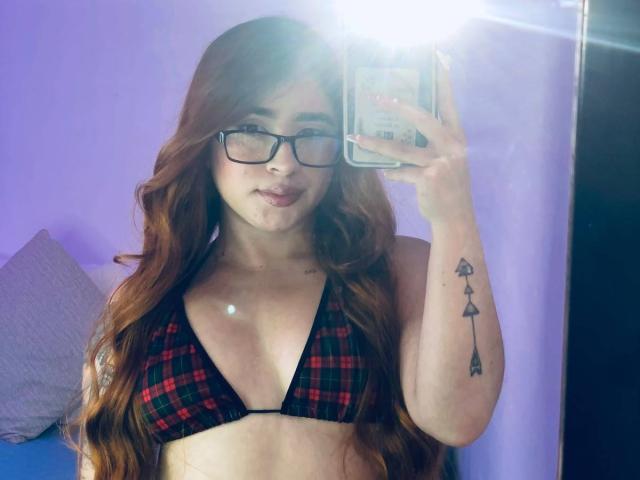 MartinaConnorr with wavy red hair, glasses, black and red plaid bikini top, and a tattoo of arrows on her right arm, taking a mirror selfie.
